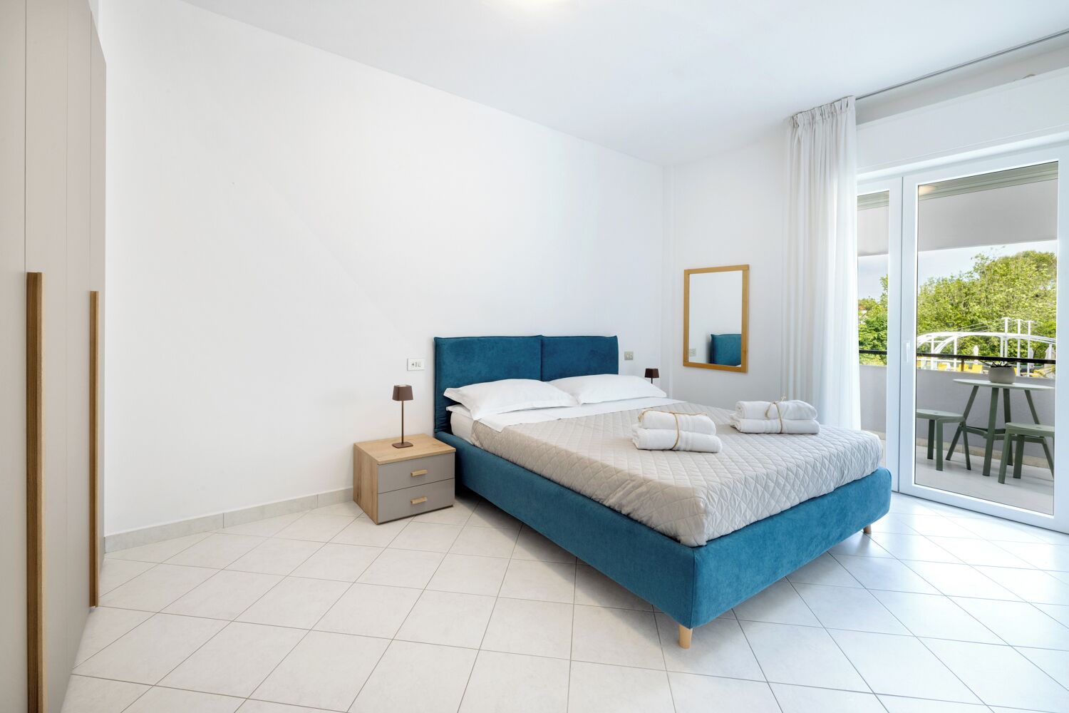 A modern bedroom with a large bed, a bedside table, a nightstand, a mirror, and a view of an outdoor area.