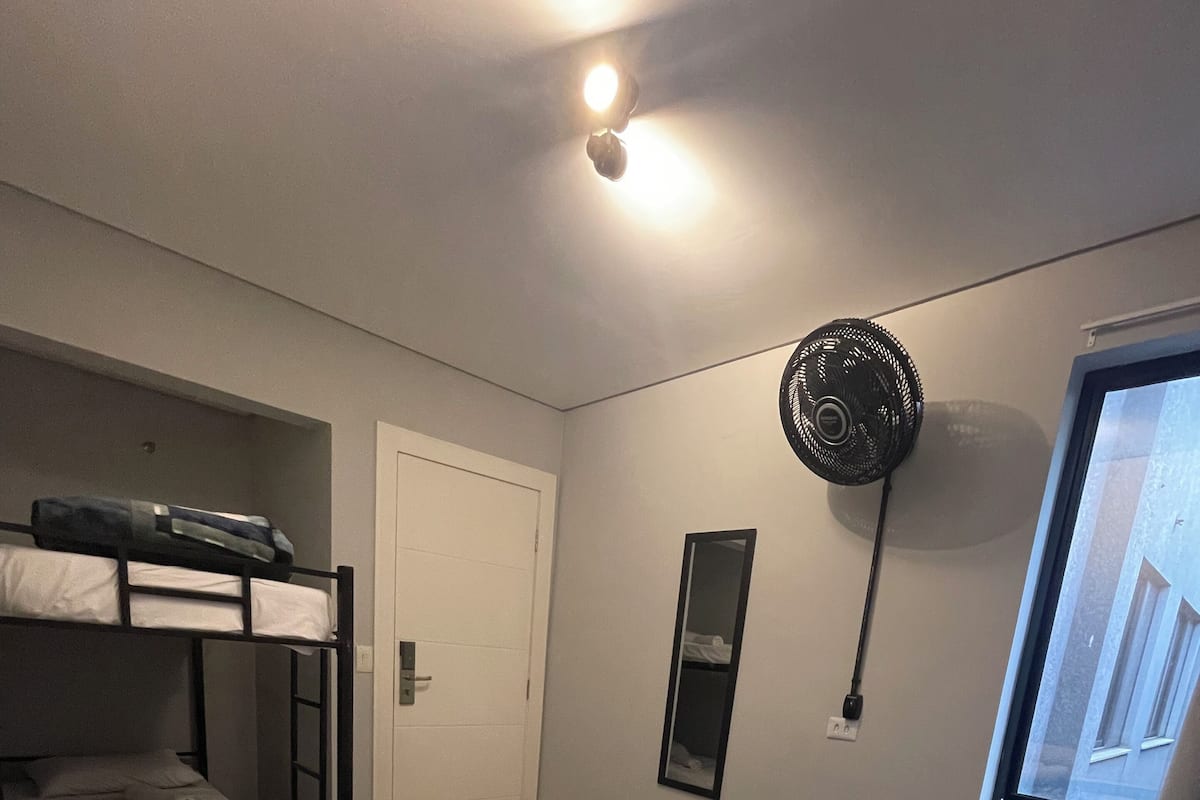 A bedroom with a bunk bed, a large bed with a patterned bedspread, a standing fan, a mirror, and a window with a view of the outside.