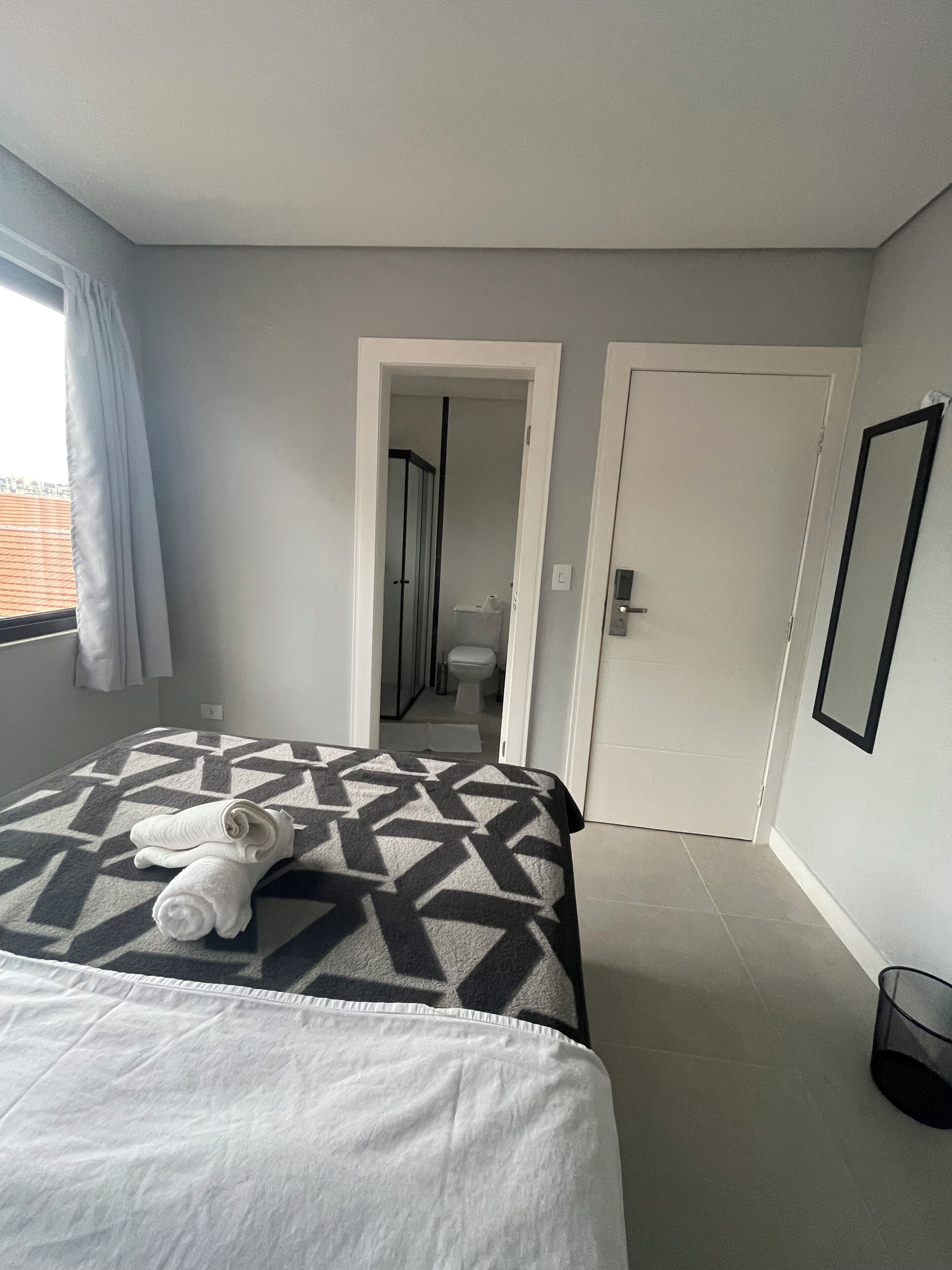 Una habitación de hotel con una cama, un baño visible a través de una puerta abierta, una ventana con cortinas y un espejo enmarcado en la pared.