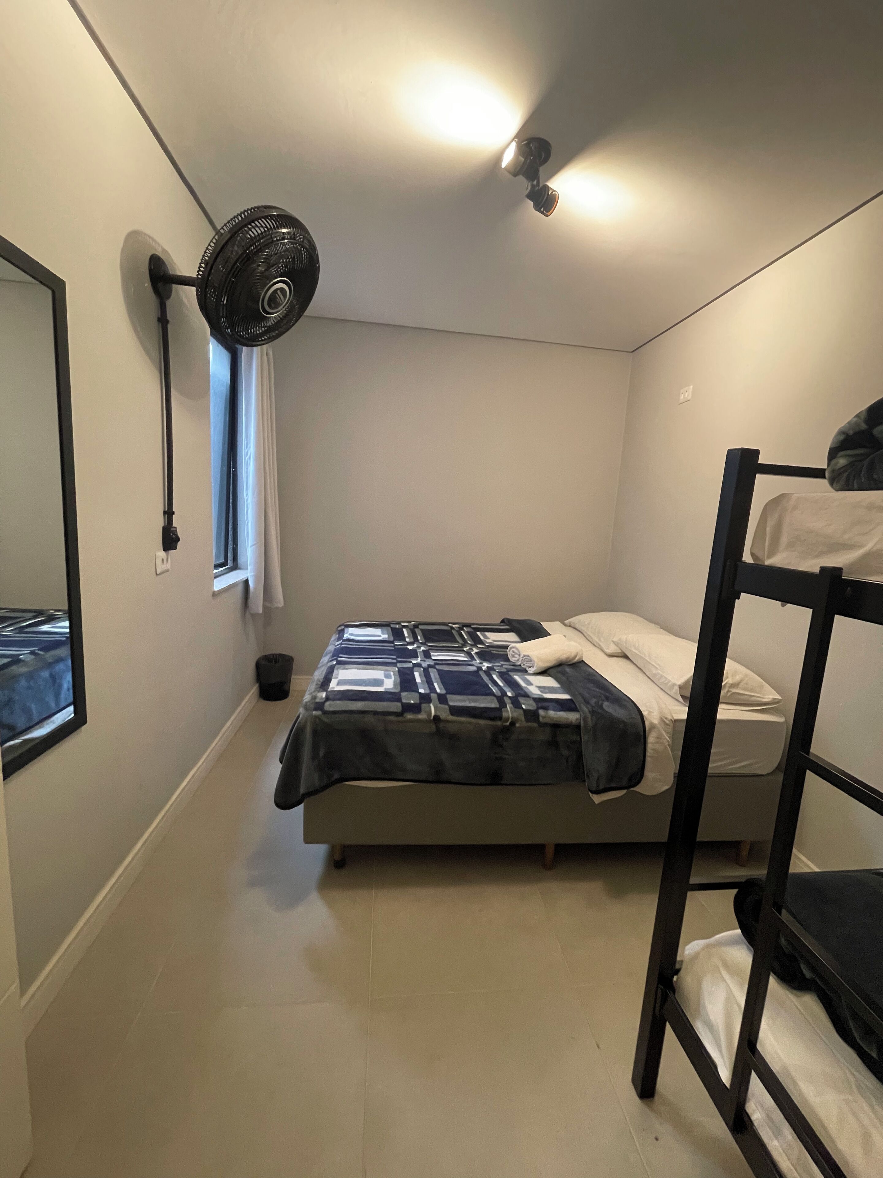 A small, single-bed room with a bunk bed on the right, a standing fan, a window with curtains, and a wall-mounted light fixture.