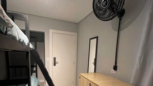 Ein Stockbett mit einer Stehleiter, eine Holzkommode, ein Standventilator, ein Spiegel und eine Tür in einem Raum mit hellen Wänden und Boden.