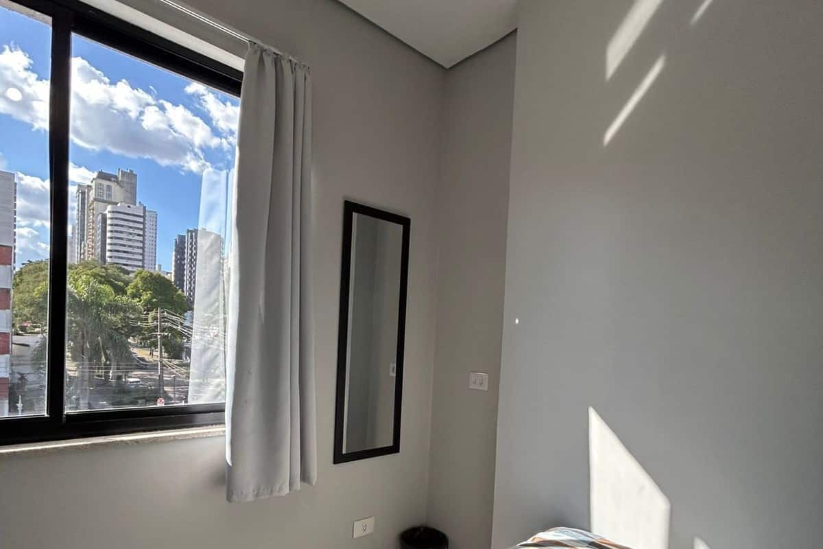 A bedroom with a bed, a window with curtains, a mirror, and a view of buildings.