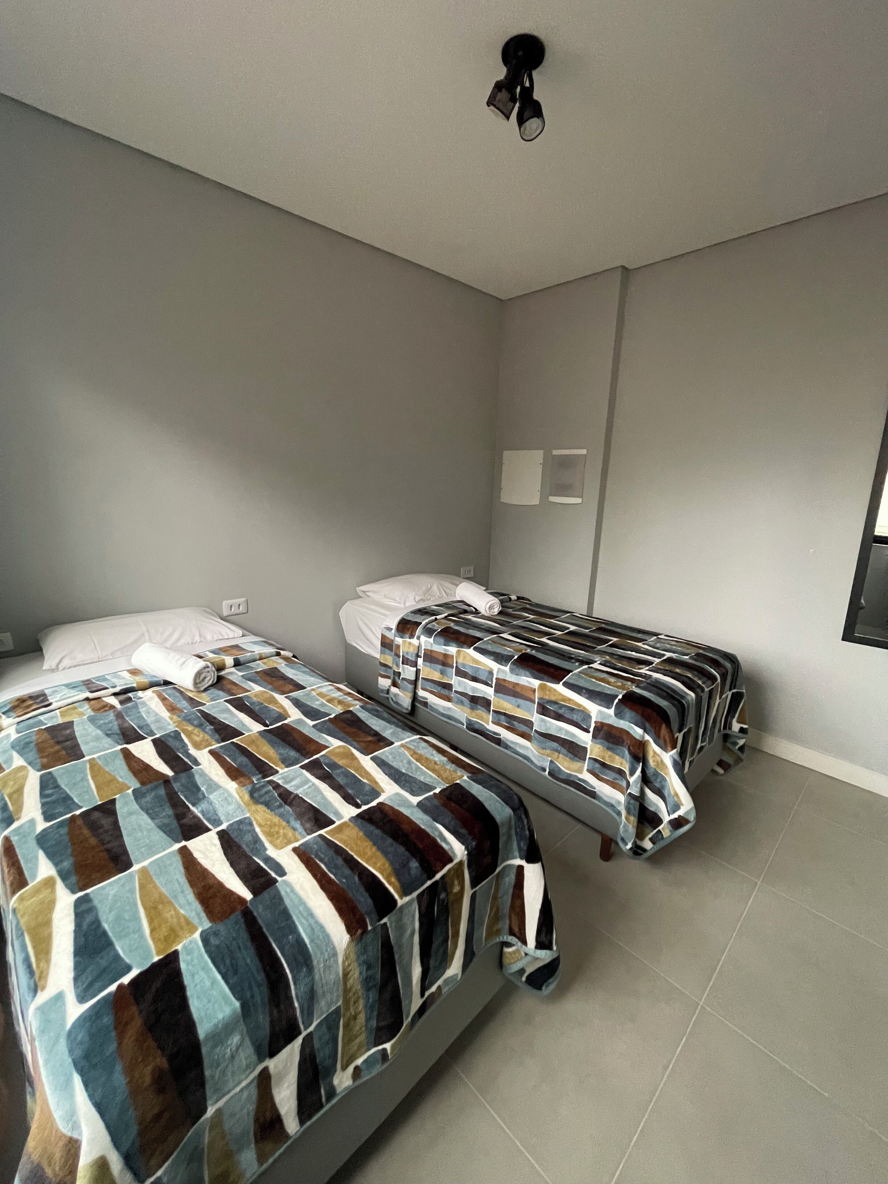 A room with two beds, each with striped bedding, a ceiling light, and a wall-mounted TV.