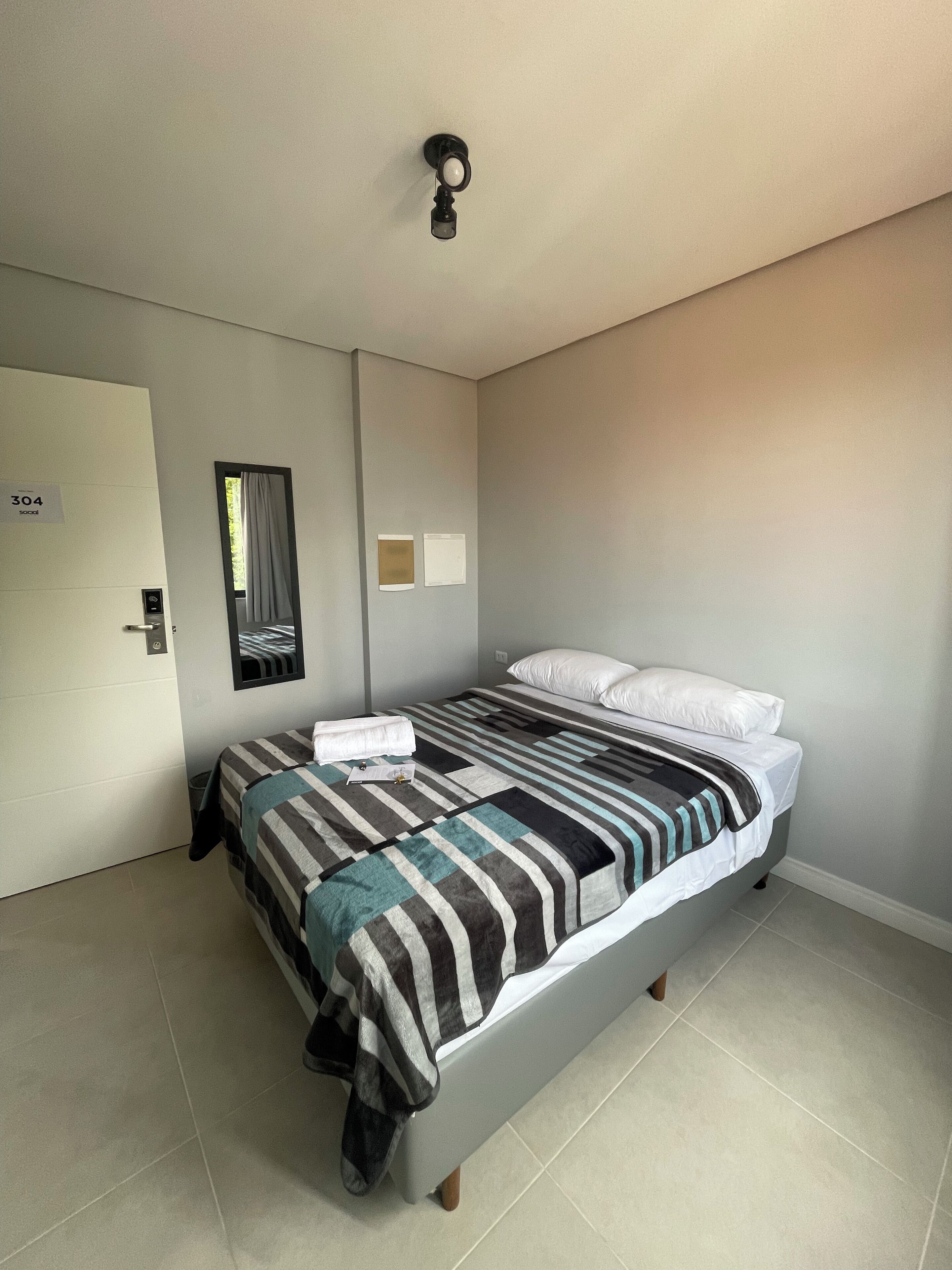 Una cama bien hecha con un edredón a rayas, una mesita de noche con una lámpara, y una puerta con un letrero con número.