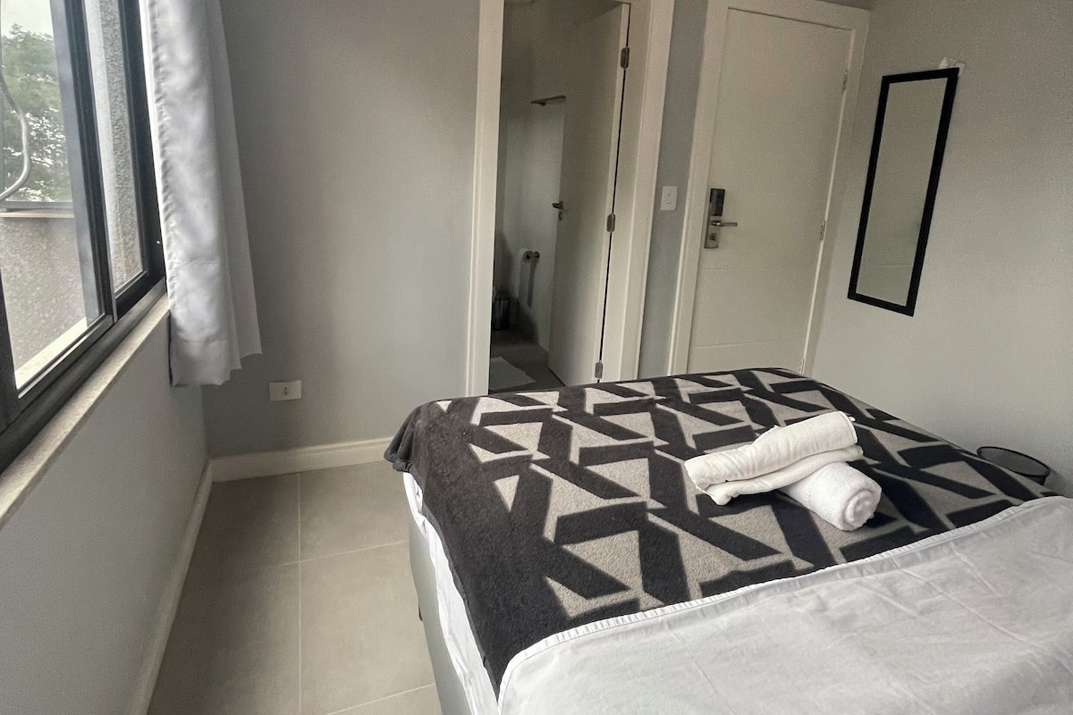 A hotel room with a bed, a window with curtains, a door, and a mirror on the wall.