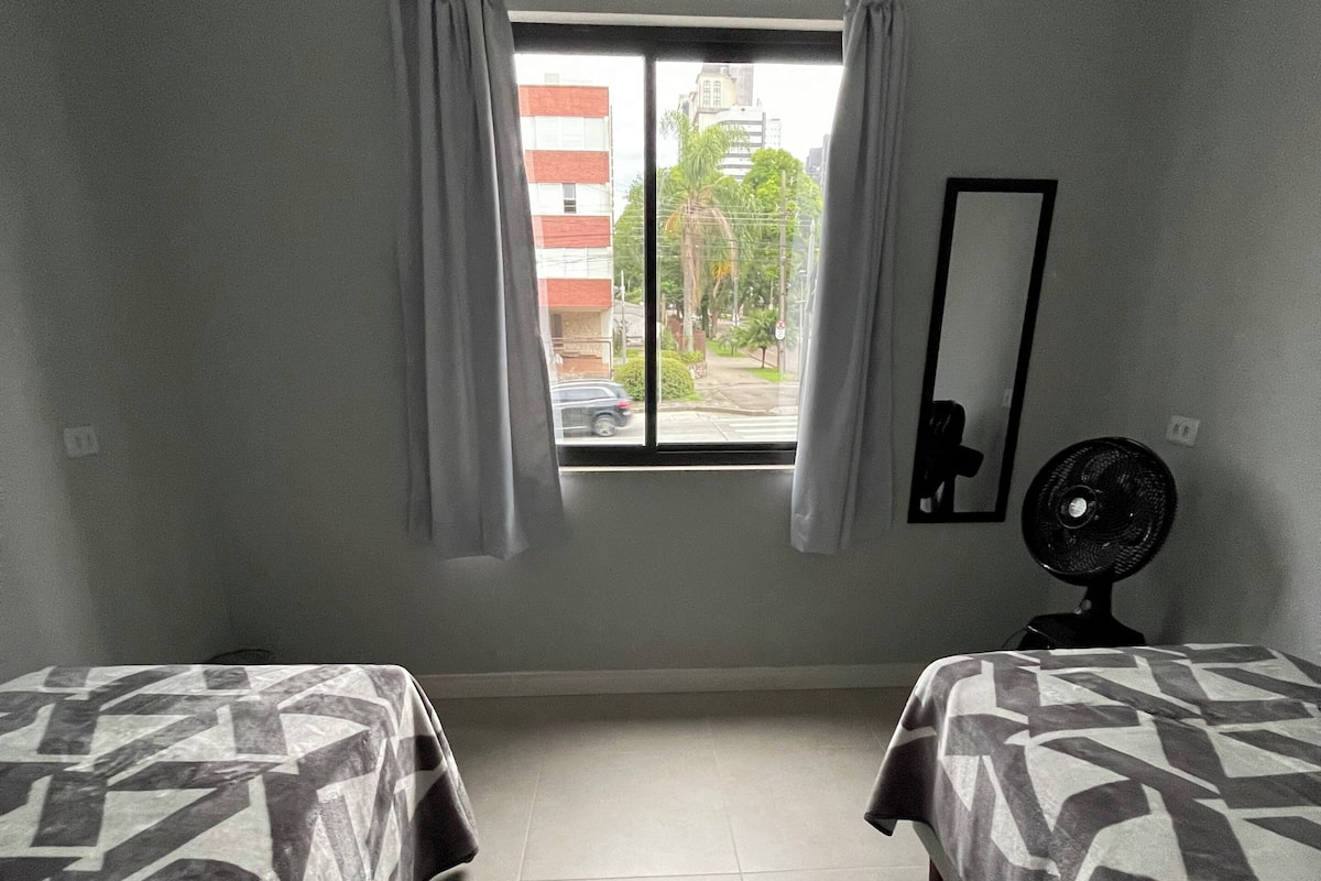 A room with two beds, a window with curtains, a standing fan, and a mirror on the wall.
