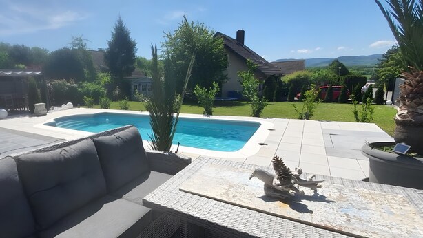 Ein Garten mit Schwimmbad, einer Terrasse mit Tisch und Kissen sowie einem Haus im Hintergrund.