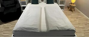 Una cama con colchón blanco y cubierta estampada en cuadrícula, una mesita de noche con lámpara y suelo de madera.