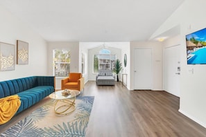 Een ruime woonkamer met een blauwe bank, een oranje fauteuil en een grijze bank. Er staat een glazen salontafel met een gele plaid.
