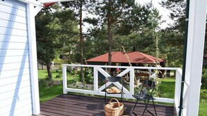 Een houten terras met een witte reling, een picknickopstelling inclusief een mand en een bord eten, en een gebouw met een rood dak op de achtergrond.