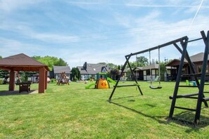 Ein Spielplatz mit Schaukeln und einer Kletterkonstruktion, umgeben von Häusern und blauem Himmel.