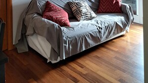 A modern living room with a grey sofa, red and patterned cushions, a wooden floor, and a large window with blinds. - Lovely and Charming Nest close to Porta Romana (Milano)