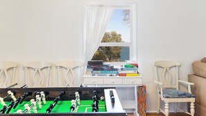 A room with a foosball table, a window with a view of trees, and a desk with books and a chair. - Treetop Tides, Sound View and Canal Front 4BR 3BA Emerald Isle! (Emerald Isle)