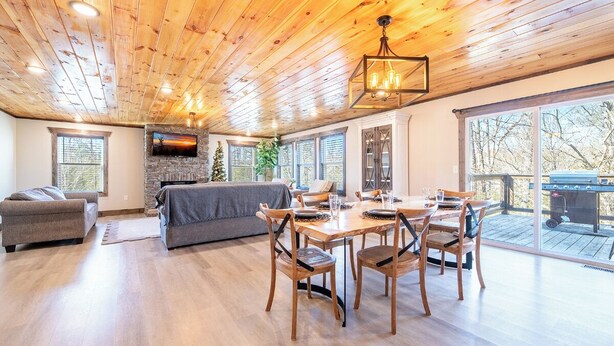 Een ruime woonkamer met een open haard, een eethoek met houten stoelen, en grote ramen met uitzicht op de omgeving.