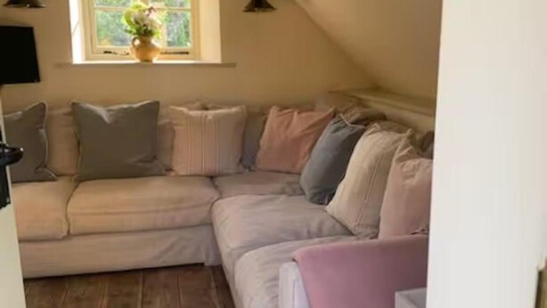 A cozy, L-shaped sectional sofa with multiple pillows in a room with wooden flooring and a window with a view of greenery. - Little Rissington, Pets, Parking, Sleeps 2 (Cheltenham)