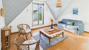 Een moderne woonkamer met een houten salontafel, rieten stoelen, een blauwe bank en een groot raam met uitzicht op groen.