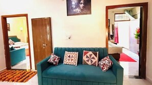 A teal sofa with patterned cushions in a room with a wooden door, a framed picture on the wall, and a view into another room with a red carpet. - Brightness in the Sky – A Cozy, Peaceful Stay with Modern Comforts (Jaipur)
