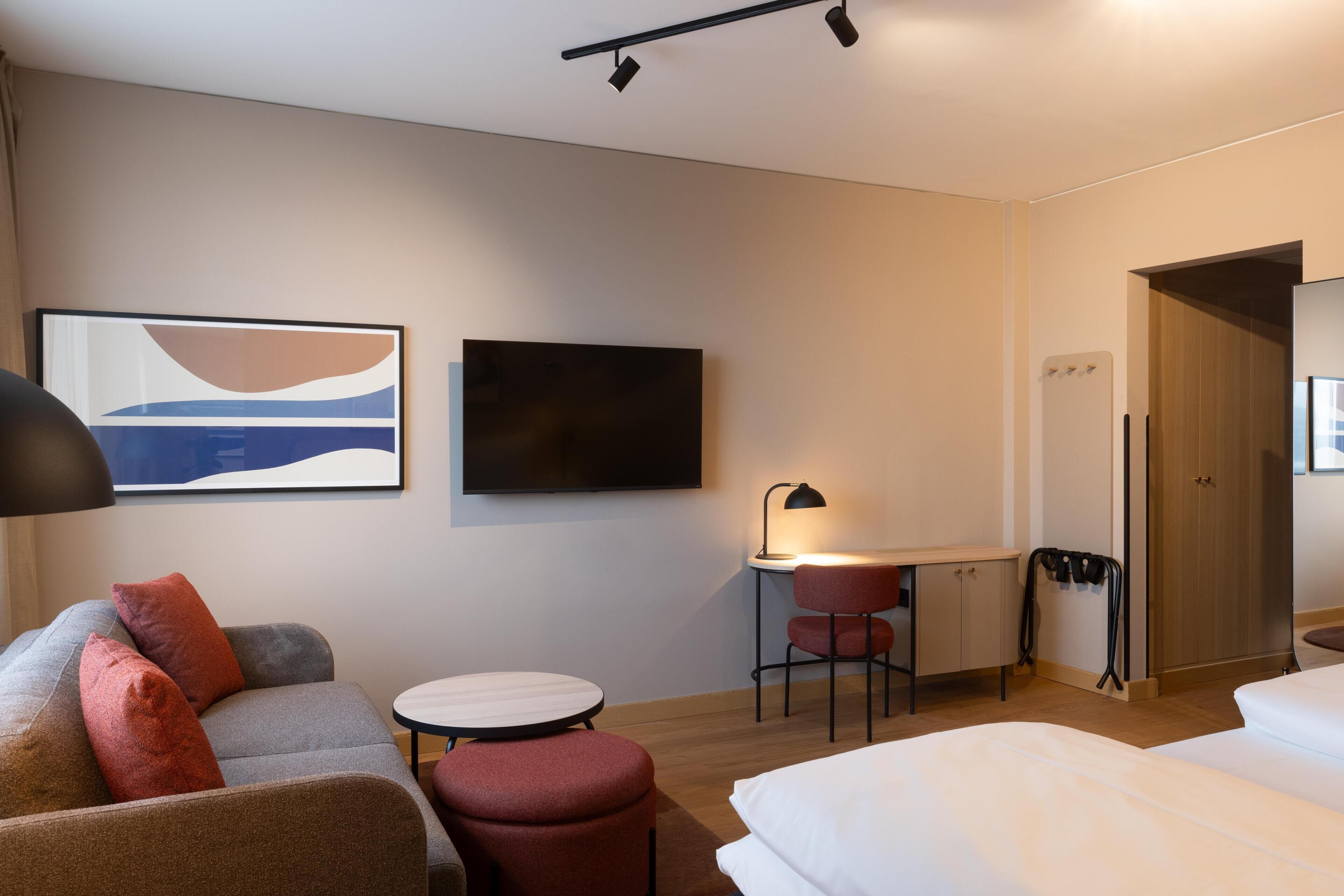 A hotel room with a bed, a sofa, a desk with a chair, a TV, and a lamp.