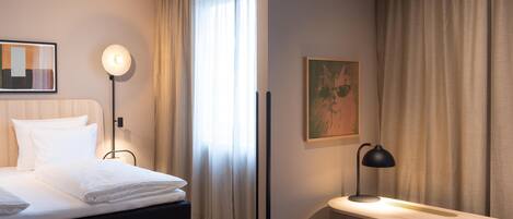 A hotel room with a bed, bedside table, desk, chair, and a painting on the wall.