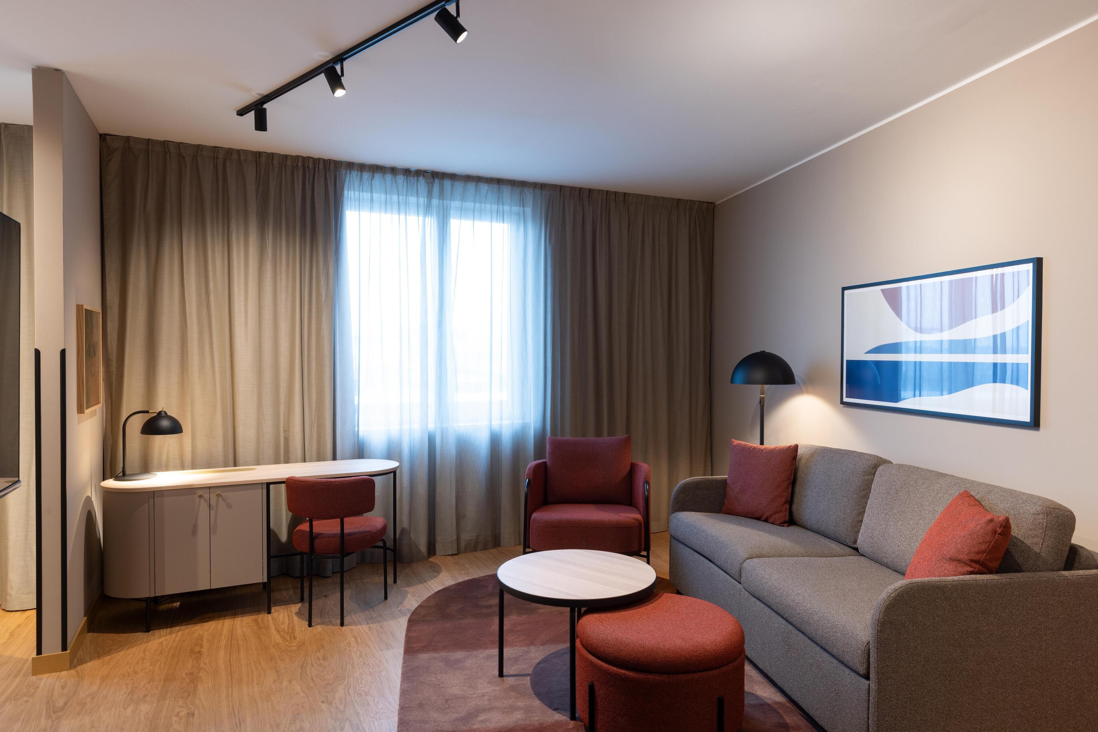 Een moderne hotelkamer met een bureau, twee stoelen, een bank, een klein voetenbankje en een lamp.