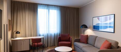 Una habitación de hotel moderna con escritorio, dos sillas, un sofá, un pequeño otoman y una lámpara.