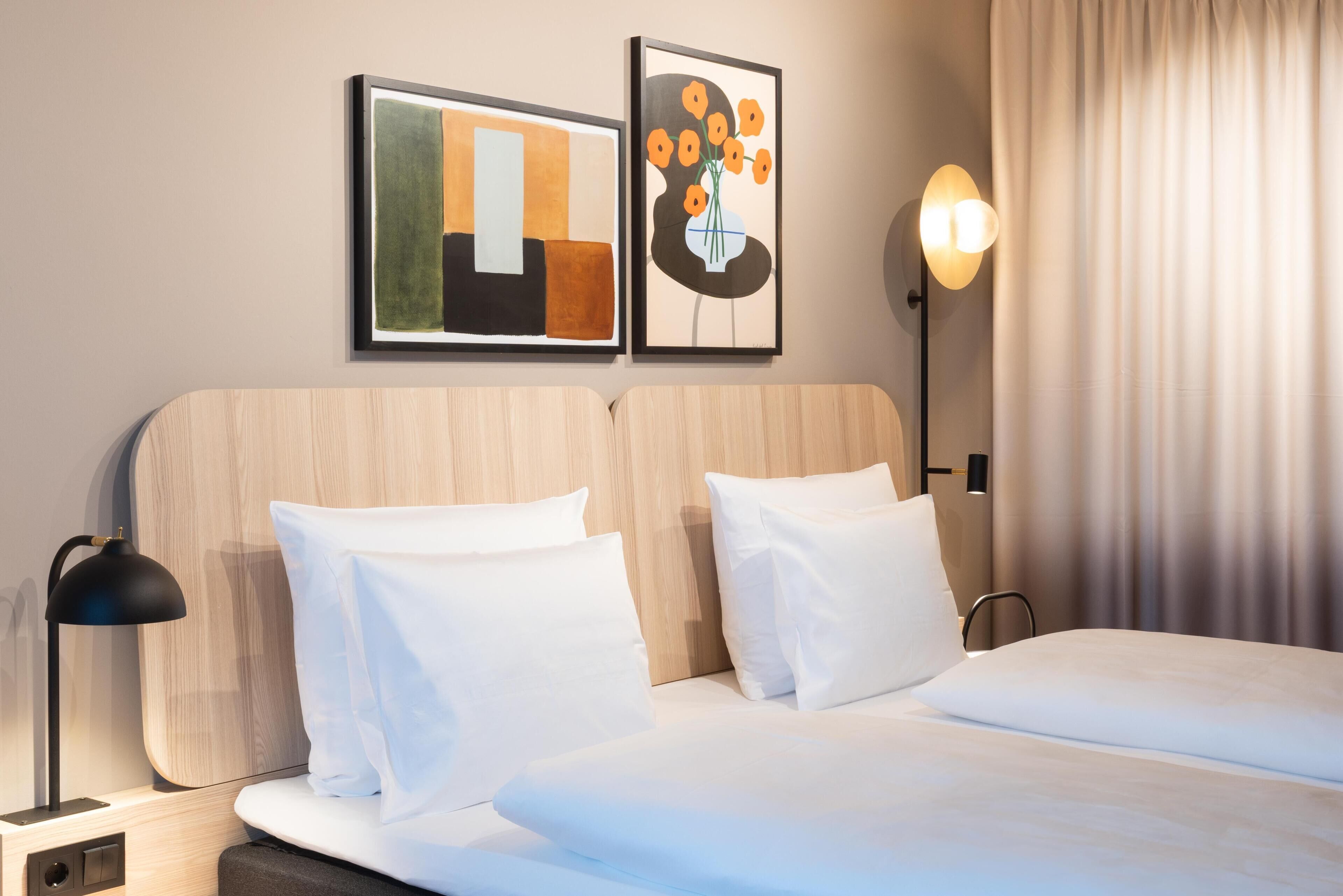 Hotellihuone, jossa on sänky, kaksi tyynyä, yöpöytä lampulla ja kaksi kehystettyä taideteosta seinällä.