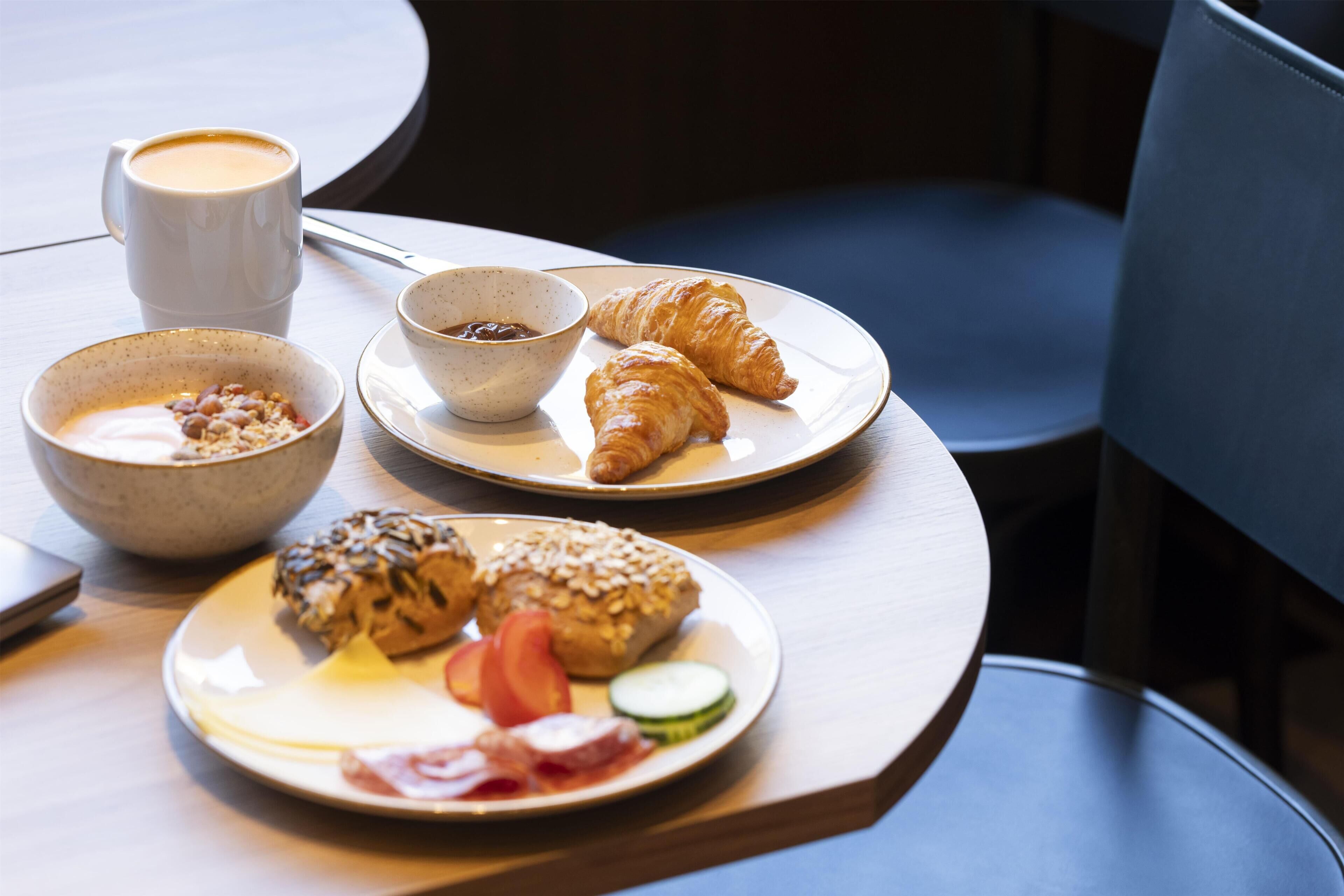 A breakfast setting with croissants, a bowl of cereal, a cup of coffee, and a plate with eggs and vegetables.