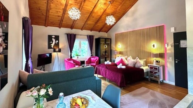 Hotelkamer met een houten plafond, een bed met rode beddengoed, een zithoek met een tafel en stoelen, en een klein bureau met een lamp.