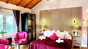 Een slaapkamer met een groot bed, een houten plafond, een nachtkastje, een stoel en een raam met gordijnen.