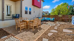 A patio with a table and chairs, a wooden deck, and a red building in the background. - Casa Tierra | Walk to Old Town via Poudre Trail (Fort Collins)