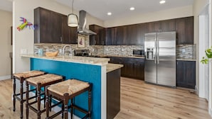 A modern kitchen with dark wood cabinets, a large island with stools, stainless steel appliances, and a patterned backsplash. - Casa Tierra | Walk to Old Town via Poudre Trail (Fort Collins)