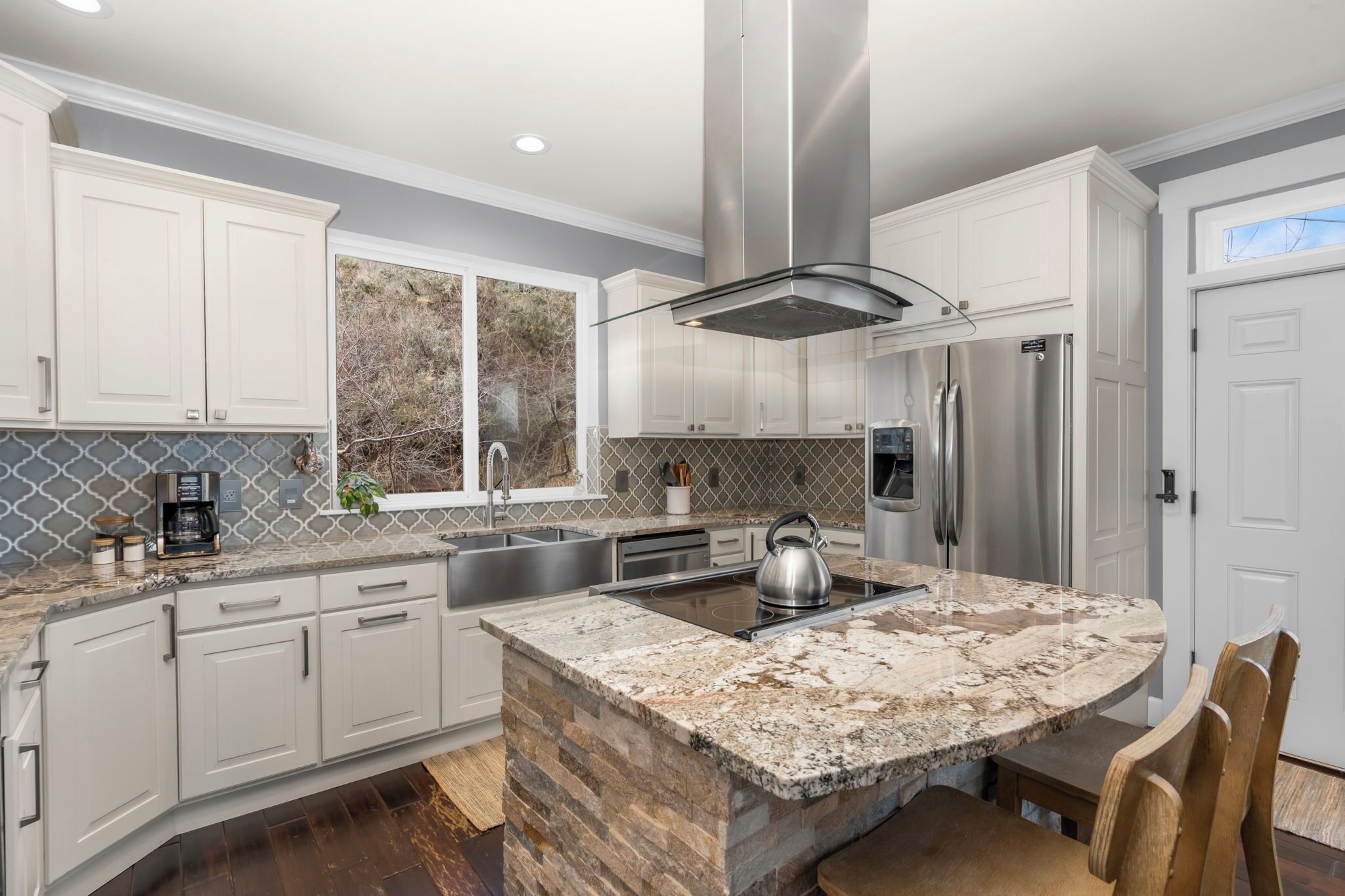 A modern kitchen with white cabinets, a granite island, stainless steel appliances, and a large window.