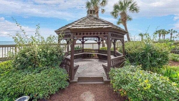 A gazebo with a wooden structure and a tiled roof, surrounded by lush greenery and palm trees, overlooking a beach. - Arcadian Dunes 11 #343- 2BR- Pool, Hot Tub (Myrtle Beach)