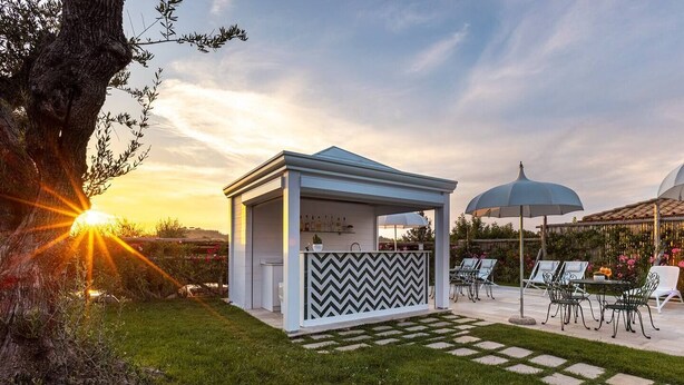 Ett utomhusområde med sittplatser i form av en pergola, komplett med bardisk, omgivet av en trädgård och en uteplats med stolar och parasoller.