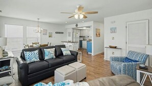 A modern living room with a black leather sofa, a blue patterned armchair, and a white ottoman. A ceiling fan and a chandelier are visible. - Ocean Edge 204- Cherry Grove 2BR- Pool (North Myrtle Beach)
