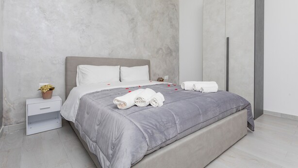 Quarto moderno com cama cinza, almofadas brancas e mesa de cabeceira com uma planta.
