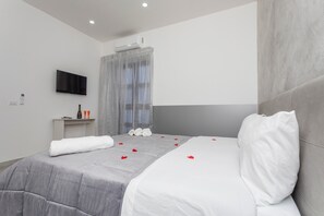 A modern hotel room with a large bed, a TV mounted on the wall, a small table with a bottle and glasses, and a shower area with a curtain. - Pm House – Double Room (Misterbianco)