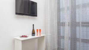 Quarto com televisão de tela plana na parede, mesa de console branca com uma garrafa e copos, e cortina de tecido leve.