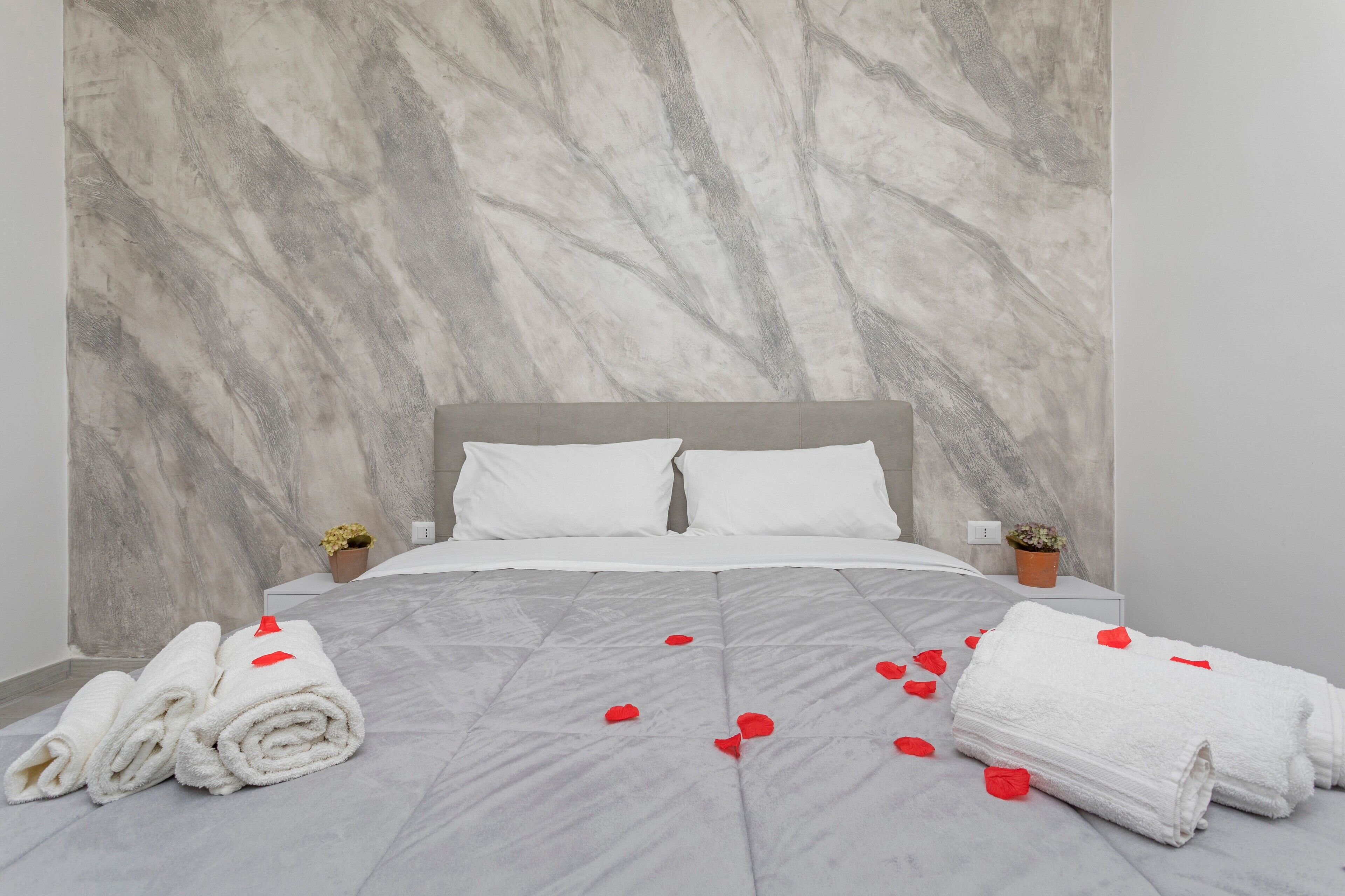 A neatly made bed with white linens and red rose petals, a marble-patterned wall, and two small potted plants on bedside tables.