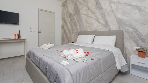 A modern hotel room with a marble wall, a bed with grey bedding, a bedside table with a plant, and a flat-screen TV mounted on the wall.