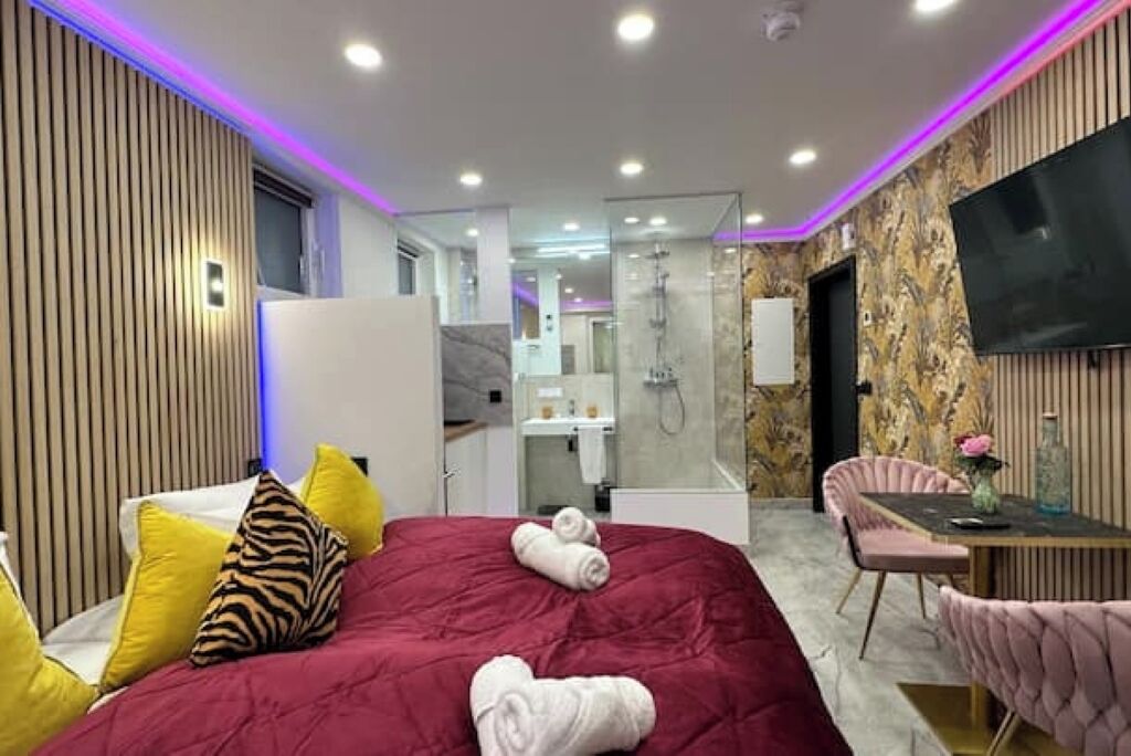 Une chambre d’hôtel moderne dotée d’un grand lit, d’un coin salon avec une table et des chaises, d’un téléviseur fixé au mur et d’un espace douche visible à travers une paroi en verre.