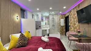 Una habitación de hotel moderna con una cama grande, una zona de estar con mesa y sillas, un televisor montado en la pared y un área de ducha visible a través de un panel de vidrio.
