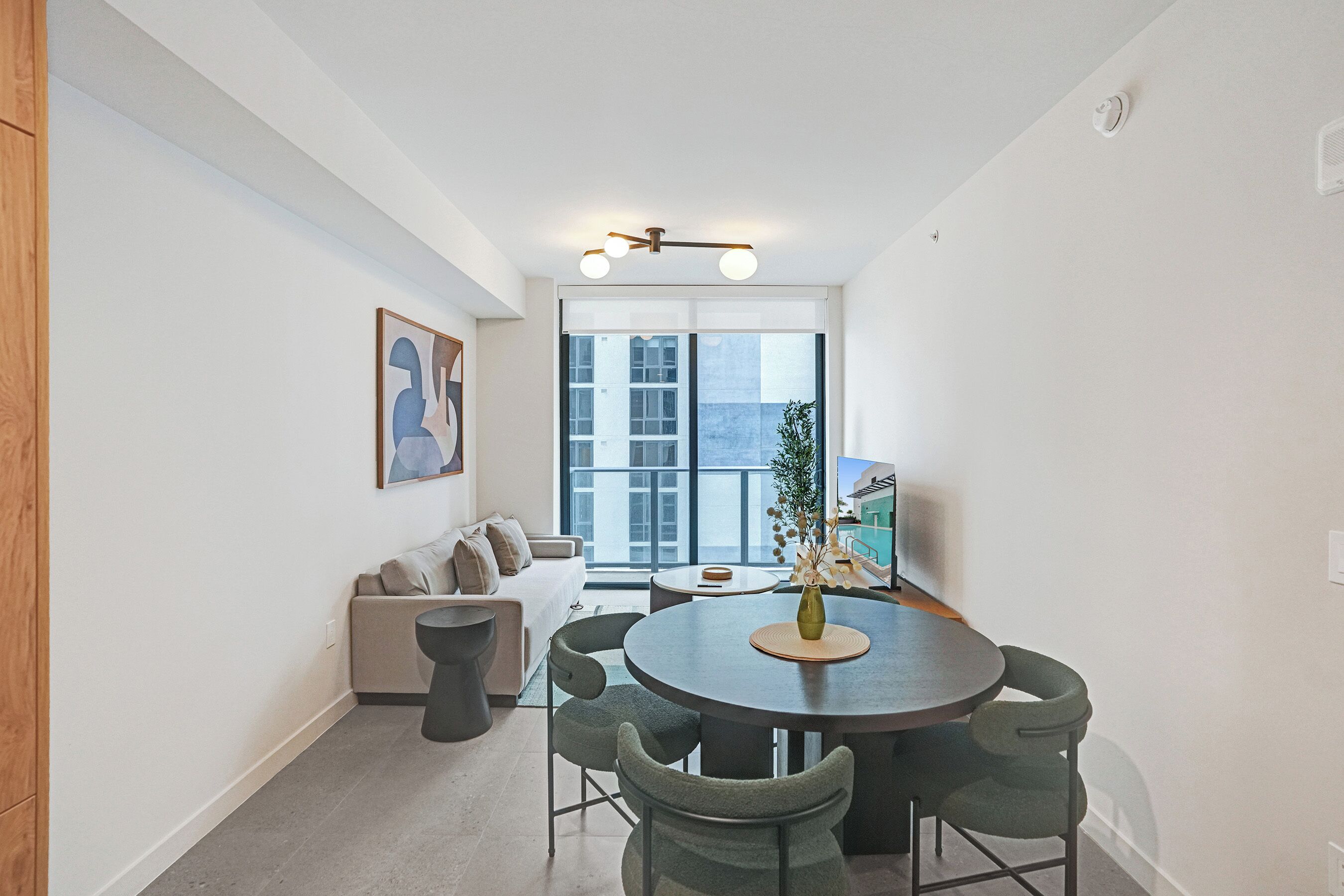 A modern living room with a round dining table, grey chairs, a sofa, and a large window with a view of buildings.