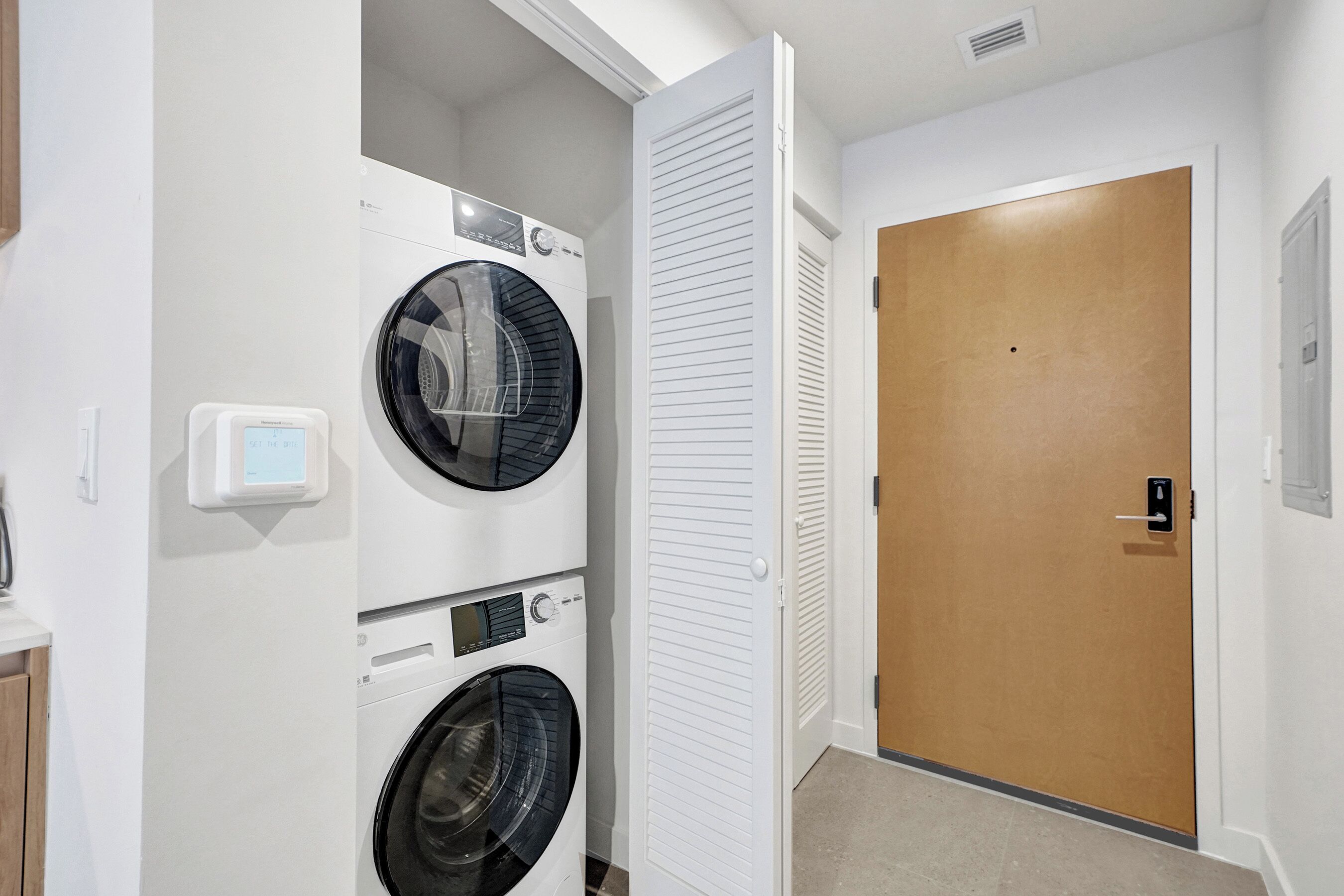 Une buanderie avec des machines à laver et des sèche-linge superposés, une porte blanche et un tapis beige.