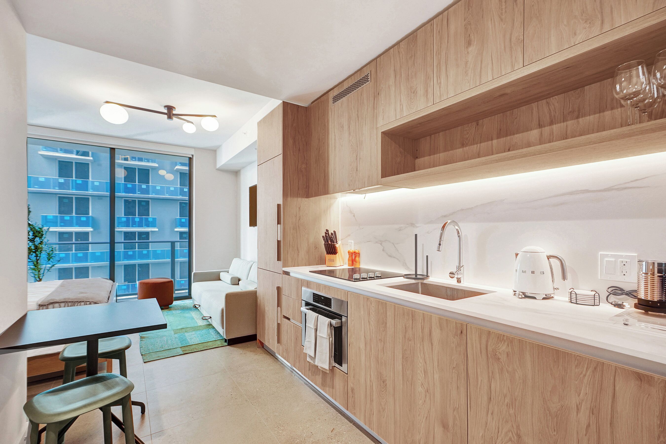 A modern kitchen with wooden cabinets, a white countertop, a built-in oven, and a large window with a view of buildings.