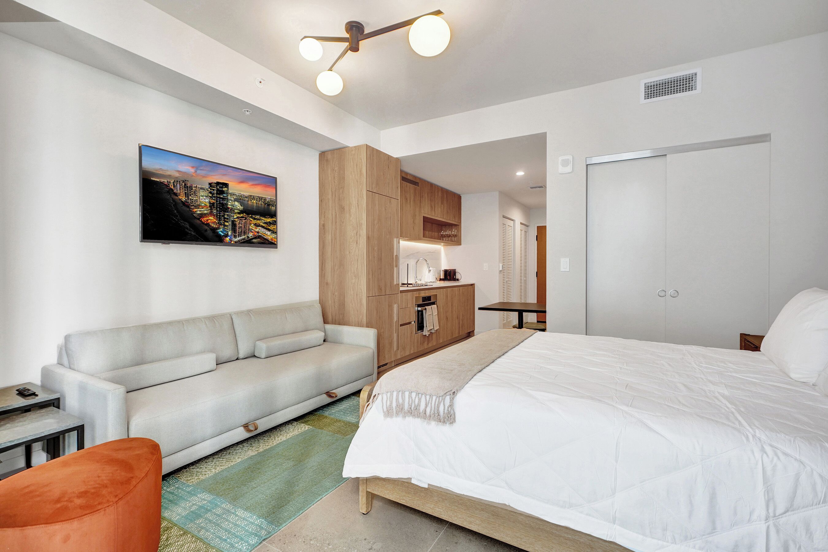 A modern hotel room with a bed, a sofa, a TV, and a kitchenette.