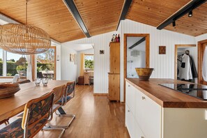 A wooden dining table with chairs, a hanging light fixture, and a kitchen counter with cabinets. - Cozy holiday home near the beach (Væggerløse)