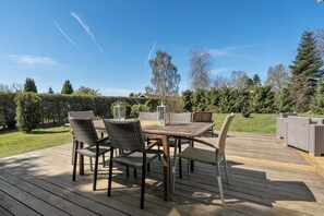An outdoor deck with a wooden table and chairs, surrounded by greenery and trees. - Cozy holiday home near the beach (Væggerløse)