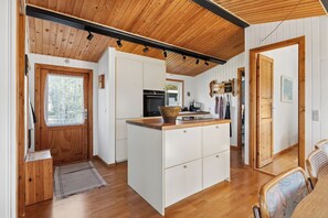 A kitchen with wooden cabinets, a central island, and a window with a lace curtain. - Cozy holiday home near the beach (Væggerløse)