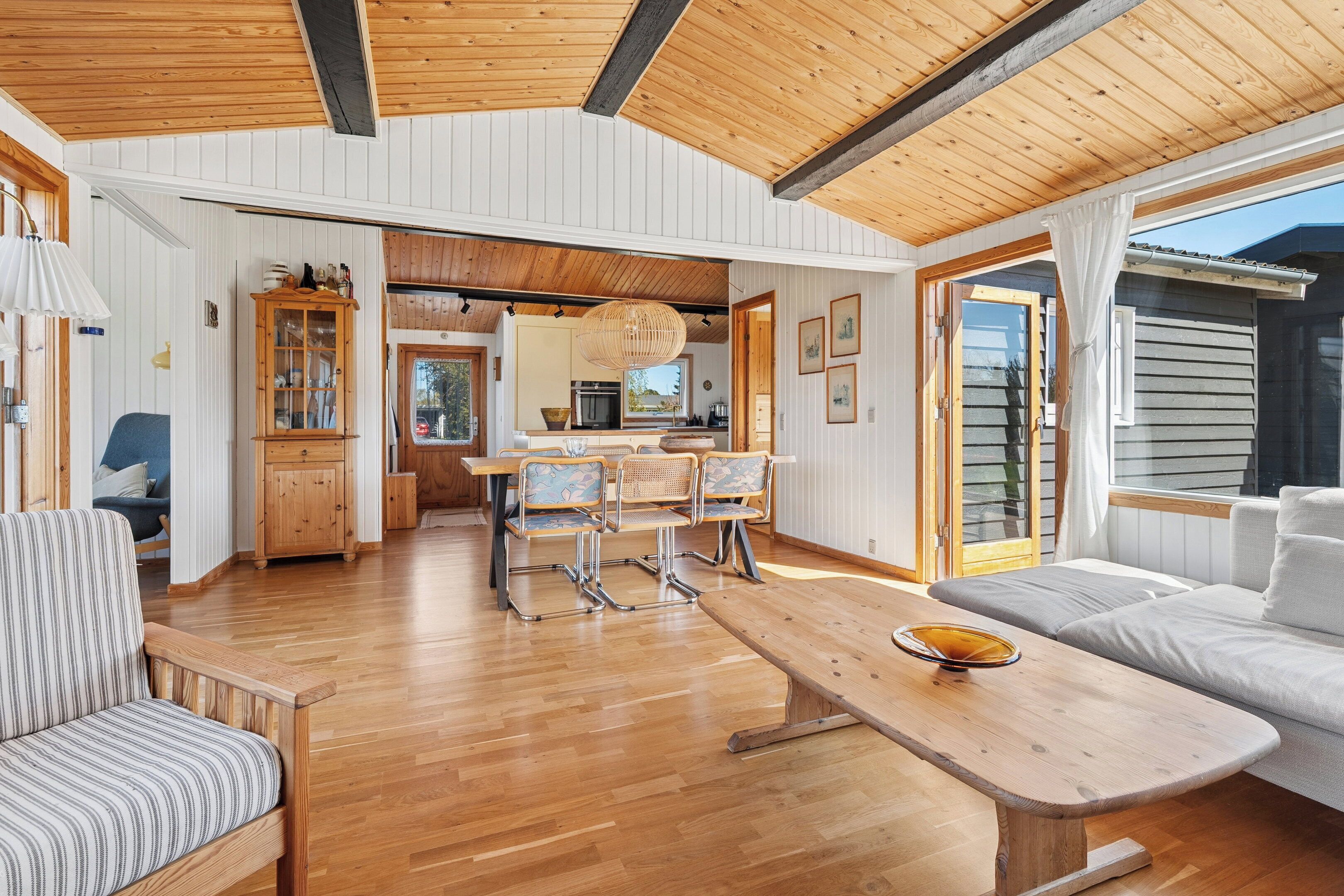Een ruime woonkamer met houten plafond, een eettafel met stoelen, een bank en een keuken op de achtergrond.